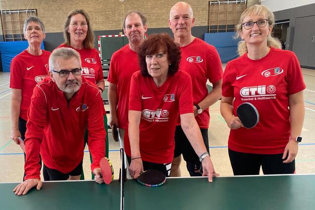 Gruppe von sieben Personen in roten T-Shirts, stehend um zwei Tischtennist&auml;fel, mit Schl&auml;gern und B&auml;llen.