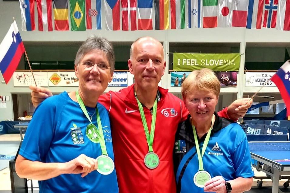 Drei Medaillengewinner posieren l&auml;chelnd mit Ihren Medaillen und Fahnen in einer Tischtennis-Halle.