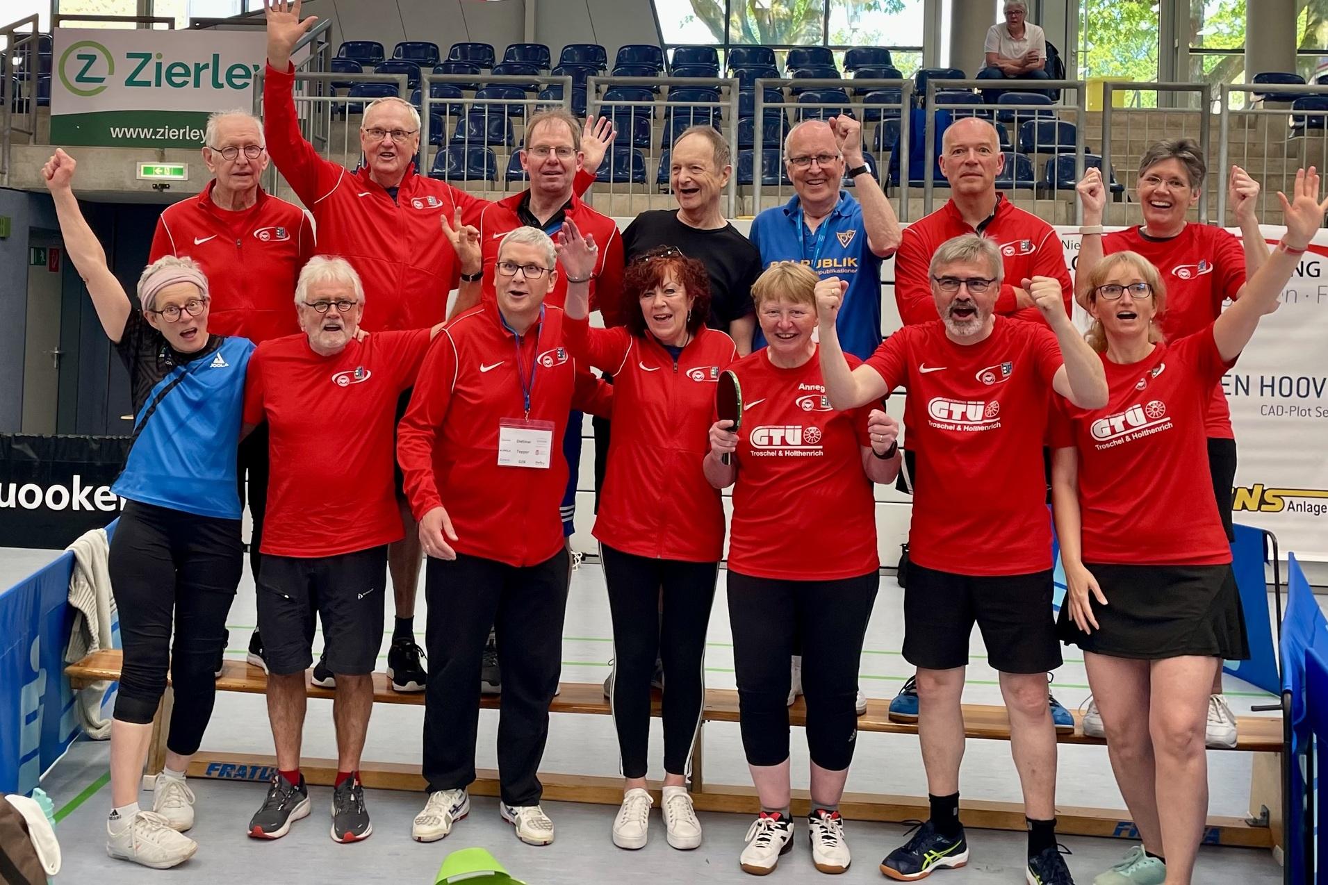 Gruppe von Tischtennisspielern in roten Shirts, die jubelnd in einer Sporthalle stehen, umgeben von Flaggen.