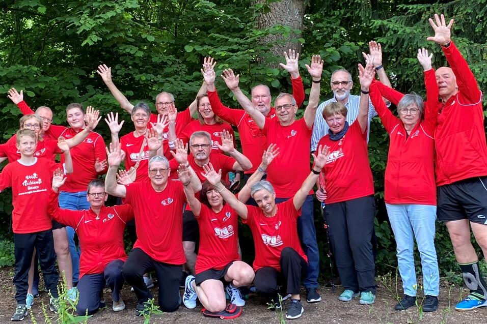 Gruppe von 16 Personen in roten T-Shirts posiert unter B&auml;umen und hebt die H&auml;nde fr&ouml;hlich in die Luft.