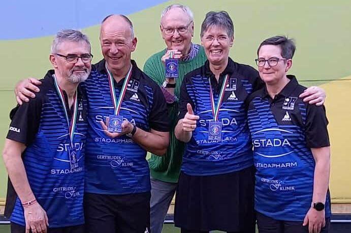 Gruppe von vier Personen in Sportkleidung, die Medaillen halten, l&auml;cheln auf einem Tischtennisplatz.
