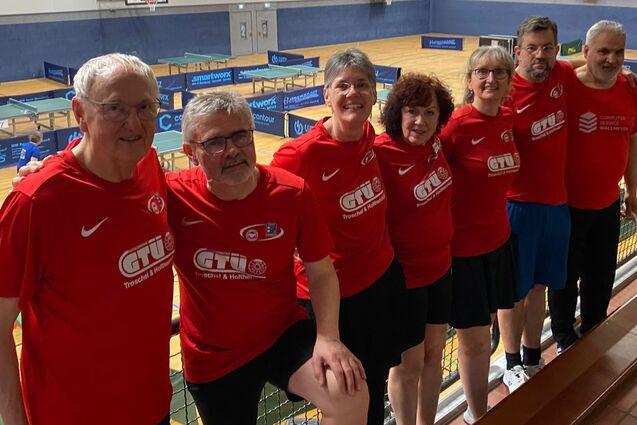 Gruppe von sieben Tischtennisspielern in roten T-Shirts l&auml;chelt auf einer Trib&uuml;ne in einer Sporthalle.