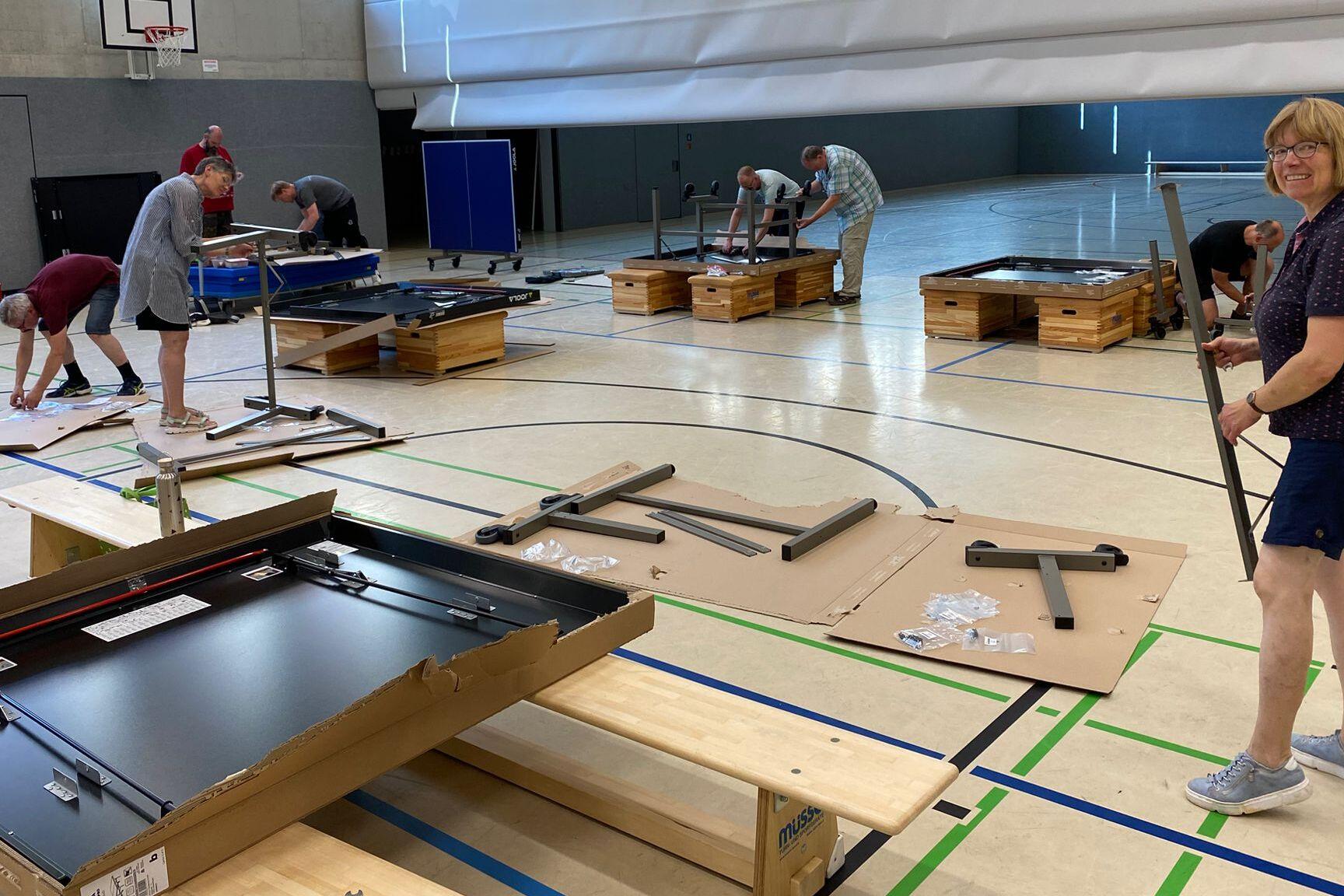 Gruppierung von Personen, die Tischtennisplatten in einer Sporthalle aufbauen, mit Materialien auf dem Boden verteilt.