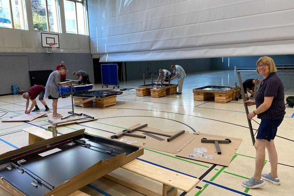 Gruppierung von Personen, die Tischtennisplatten in einer Sporthalle aufbauen, mit Materialien auf dem Boden verteilt.