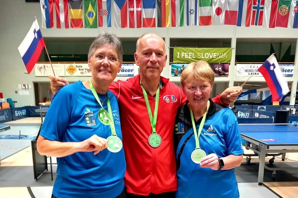 Drei Medaillengewinner posieren lächelnd mit Ihren Medaillen und Fahnen in einer Tischtennis-Halle.