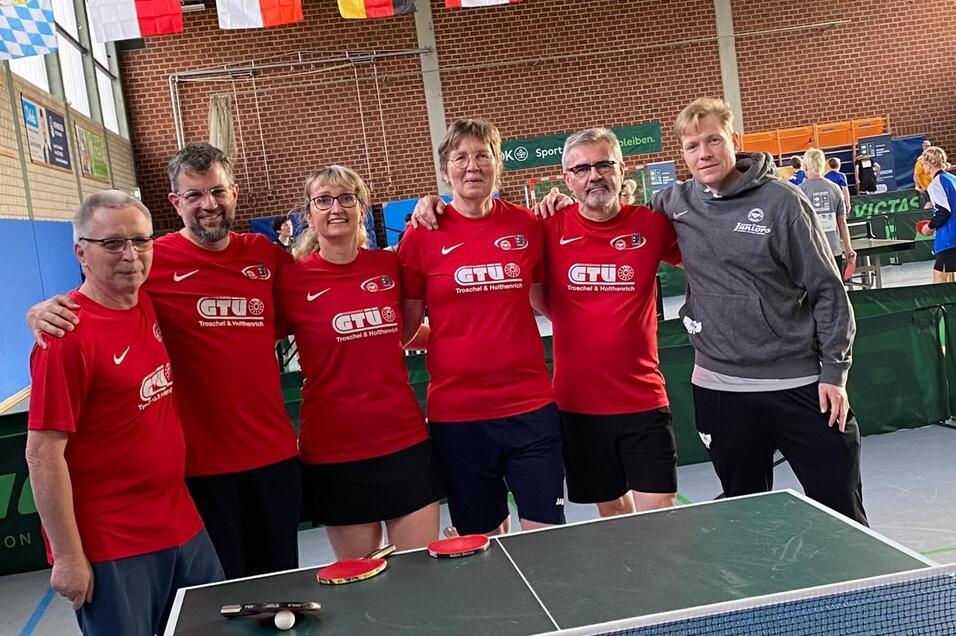 Gruppe von f&uuml;nf Tischtennisspielern in roten T-Shirts posiert neben einem Tischtennistisch in einer Halle.
