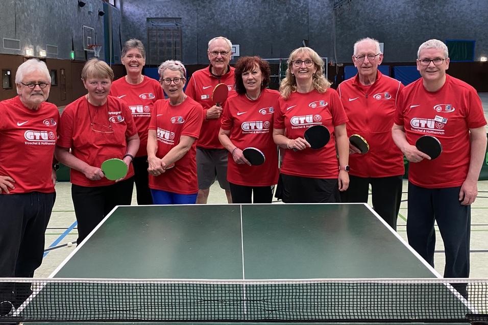 Gruppe von zehn Personen in roten T-Shirts, die Tischtennisschl&auml;ger in einer Halle halten, mit Tischtennisplatte im Vordergrund.