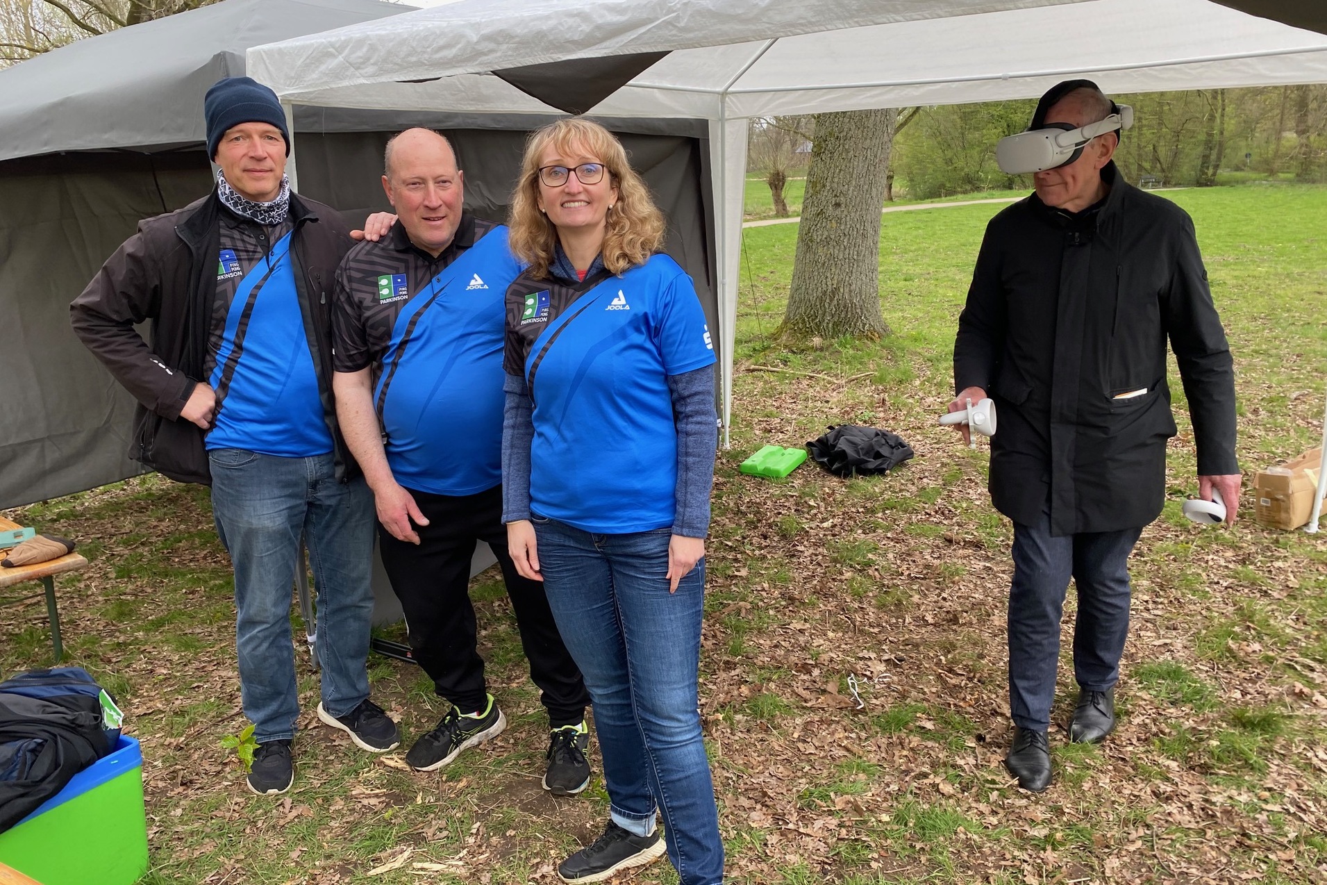 Vier Personen in blauen Shirts stehen unter einem Zelt; eine Person tr&auml;gt eine VR-Brille und schaut weg.