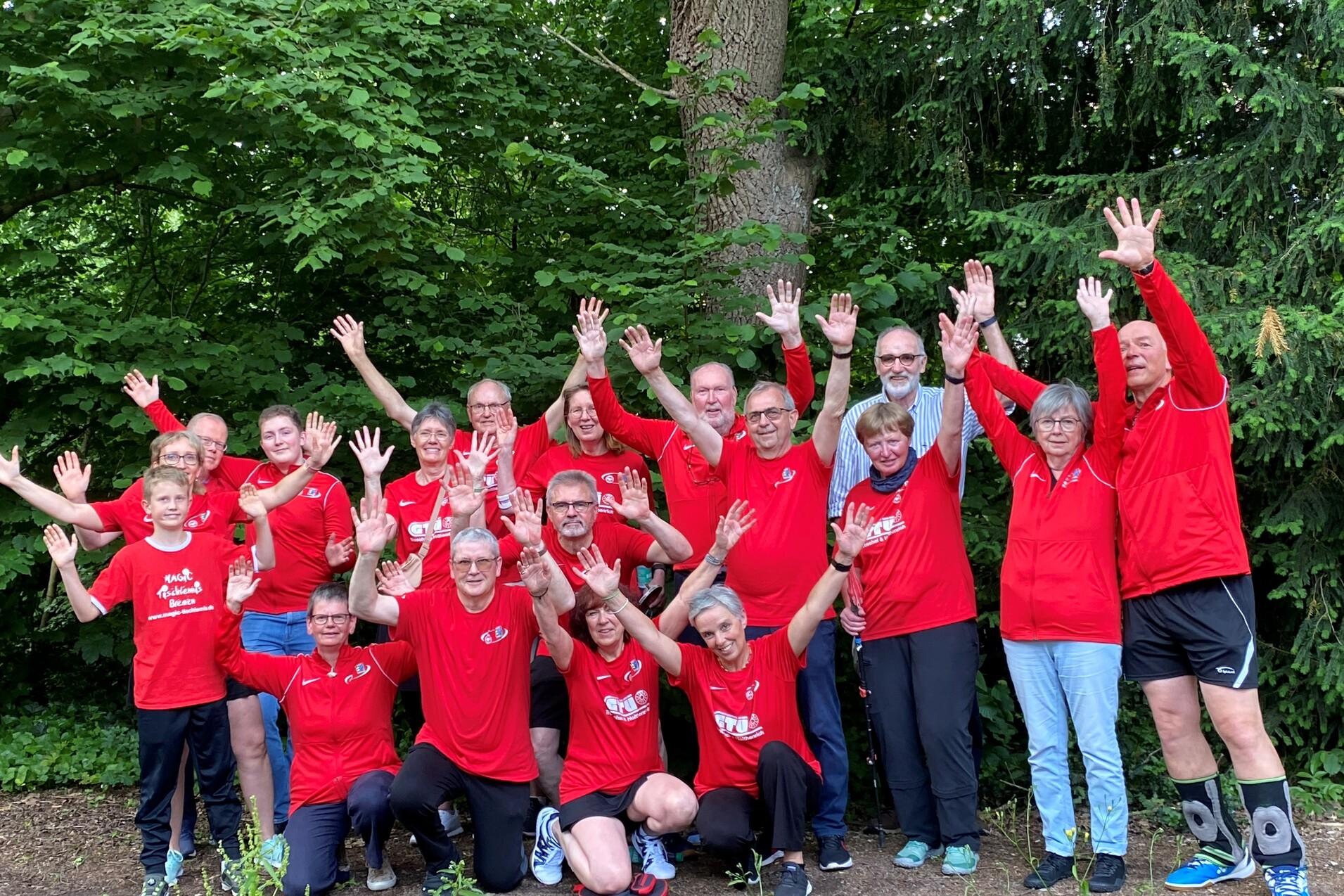 Gruppe von 16 Personen in roten T-Shirts posiert unter Bäumen und hebt die Hände fröhlich in die Luft.