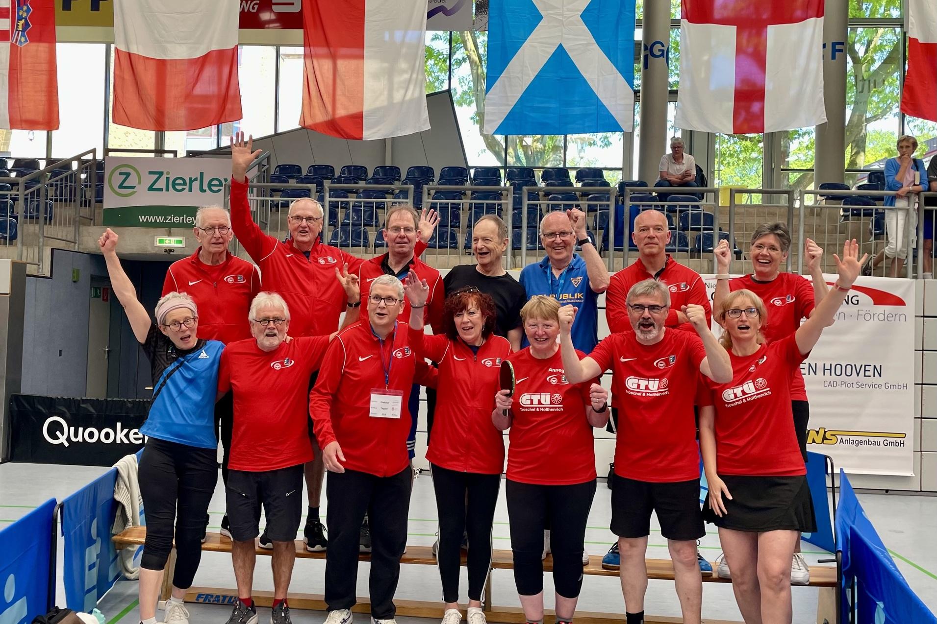 Gruppe von Tischtennisspielern in roten Shirts, die jubelnd in einer Sporthalle stehen, umgeben von Flaggen.