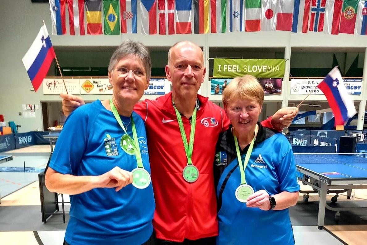 Drei Medaillengewinner posieren lächelnd mit Ihren Medaillen und Fahnen in einer Tischtennis-Halle.