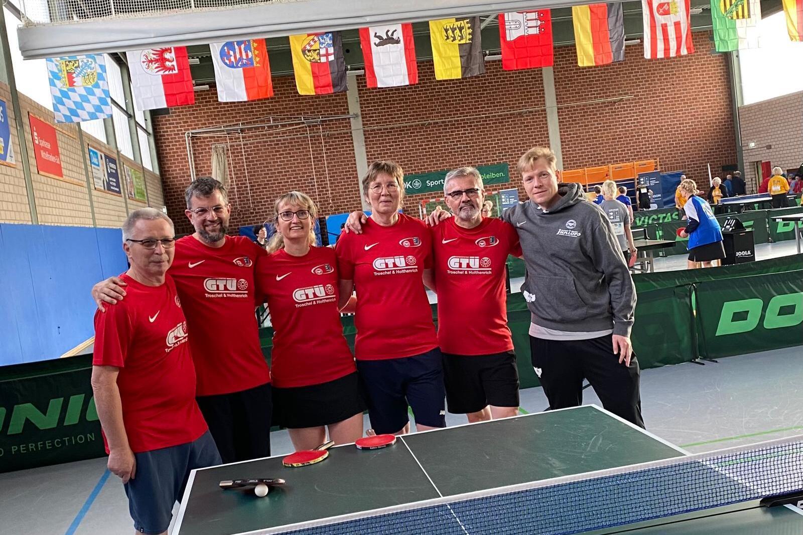 Gruppe von fünf Tischtennisspielern in roten T-Shirts posiert neben einem Tischtennistisch in einer Halle.