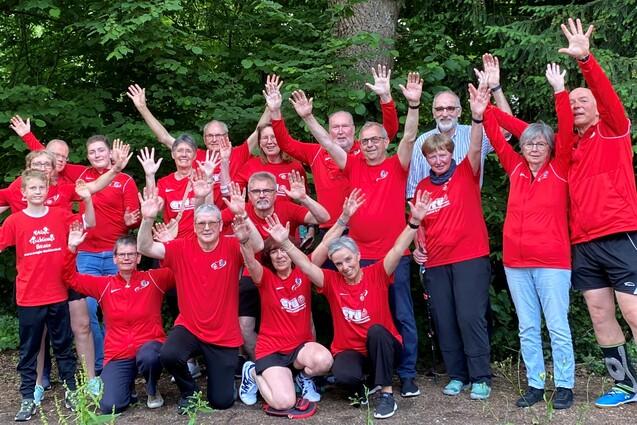 Gruppe von 16 Personen in roten T-Shirts posiert unter B&auml;umen und hebt die H&auml;nde fr&ouml;hlich in die Luft.