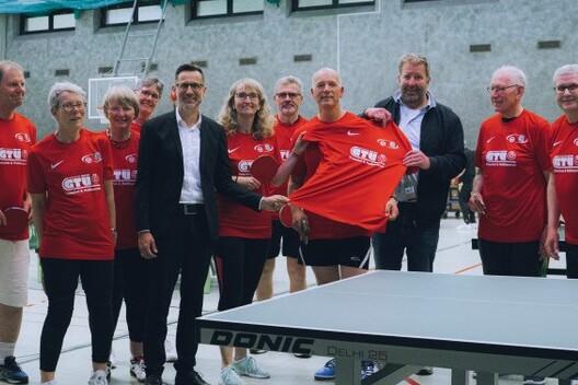 Gruppe von zw&ouml;lf Personen in roten T-Shirts stehen neben einem Tischtennistisch in einer Sporthalle.