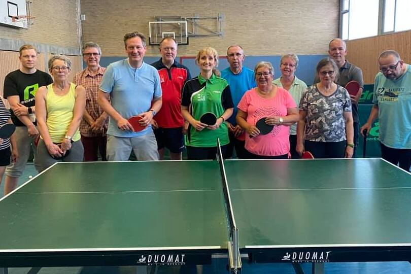 Gruppe von zw&ouml;lf Personen mit Tischtennisschl&auml;gern vor einem Tischtennistisch in einer Sporthalle.
