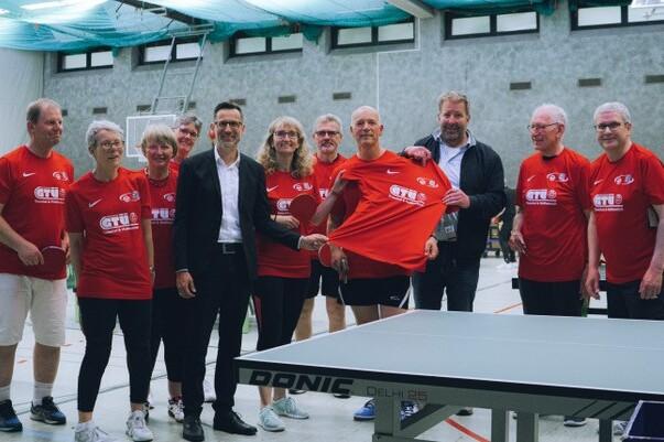 Gruppe von zw&ouml;lf Personen in roten T-Shirts stehen neben einem Tischtennistisch in einer Sporthalle.
