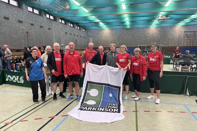 Gruppe von Tischtennisspielern in rot und blau mit Banner "Ping Pong Parkinson" in einer Sporthalle.