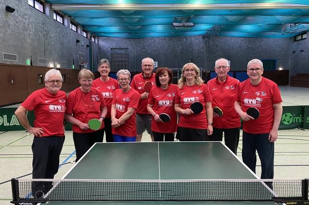 Gruppe von zehn Personen in roten T-Shirts, die Tischtennisschl&auml;ger in einer Halle halten, mit Tischtennisplatte im Vordergrund.