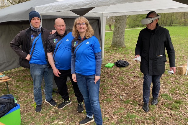 Vier Personen in blauen Shirts stehen unter einem Zelt; eine Person tr&auml;gt eine VR-Brille und schaut weg.