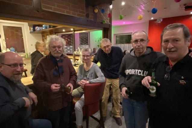 Gruppe von f&uuml;nf Personen steht in einer Bar, einige halten Getr&auml;nke, bunte Dekoration h&auml;ngt an der Decke.