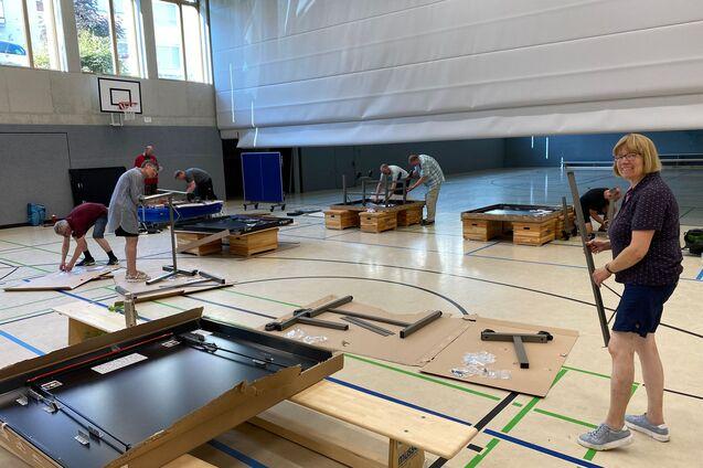 Gruppierung von Personen, die Tischtennisplatten in einer Sporthalle aufbauen, mit Materialien auf dem Boden verteilt.