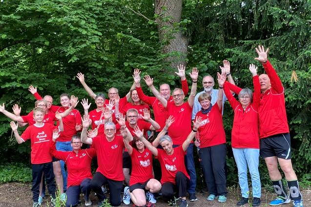 Gruppe von 16 Personen in roten T-Shirts posiert unter B&auml;umen und hebt die H&auml;nde fr&ouml;hlich in die Luft.