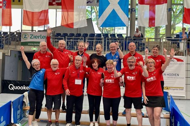Gruppe von Tischtennisspielern in roten Shirts, die jubelnd in einer Sporthalle stehen, umgeben von Flaggen.