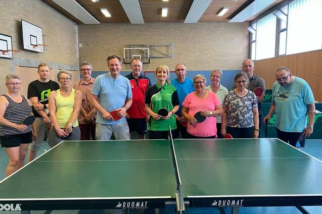 Gruppe von zw&ouml;lf Personen mit Tischtennisschl&auml;gern vor einem Tischtennistisch in einer Sporthalle.