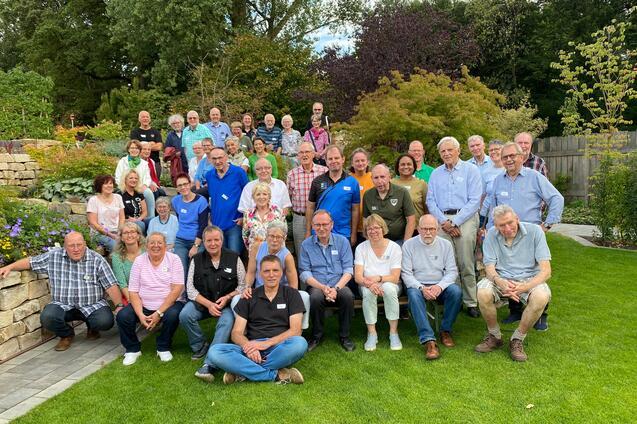 Gruppenfoto von etwa 40 Personen im Garten, umgeben von Pflanzen und B&auml;umen, l&auml;chelnd und in festlicher Stimmung.