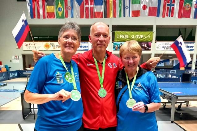Drei Medaillengewinner posieren l&auml;chelnd mit Ihren Medaillen und Fahnen in einer Tischtennis-Halle.