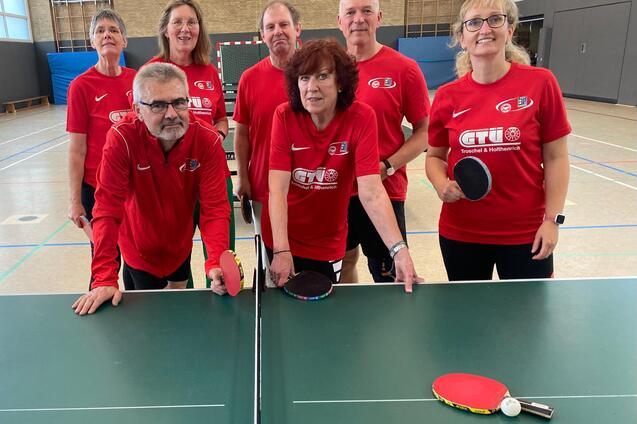 Gruppe von sieben Personen in roten T-Shirts, stehend um zwei Tischtennist&auml;fel, mit Schl&auml;gern und B&auml;llen.