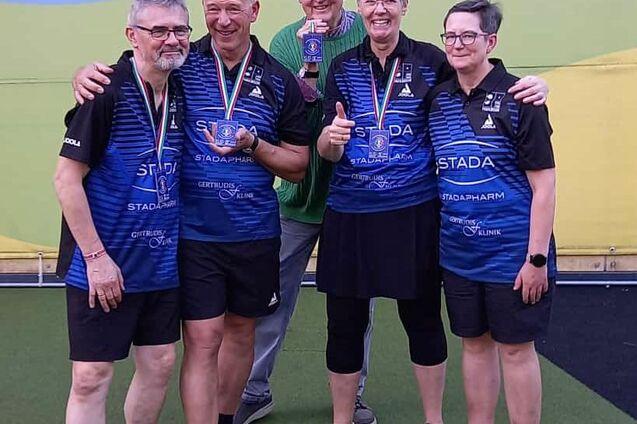 Gruppe von vier Personen in Sportkleidung, die Medaillen halten, l&auml;cheln auf einem Tischtennisplatz.