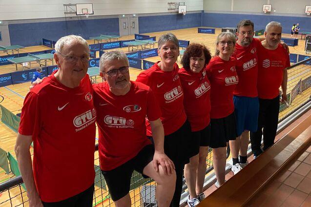 Gruppe von sieben Tischtennisspielern in roten T-Shirts l&auml;chelt auf einer Trib&uuml;ne in einer Sporthalle.