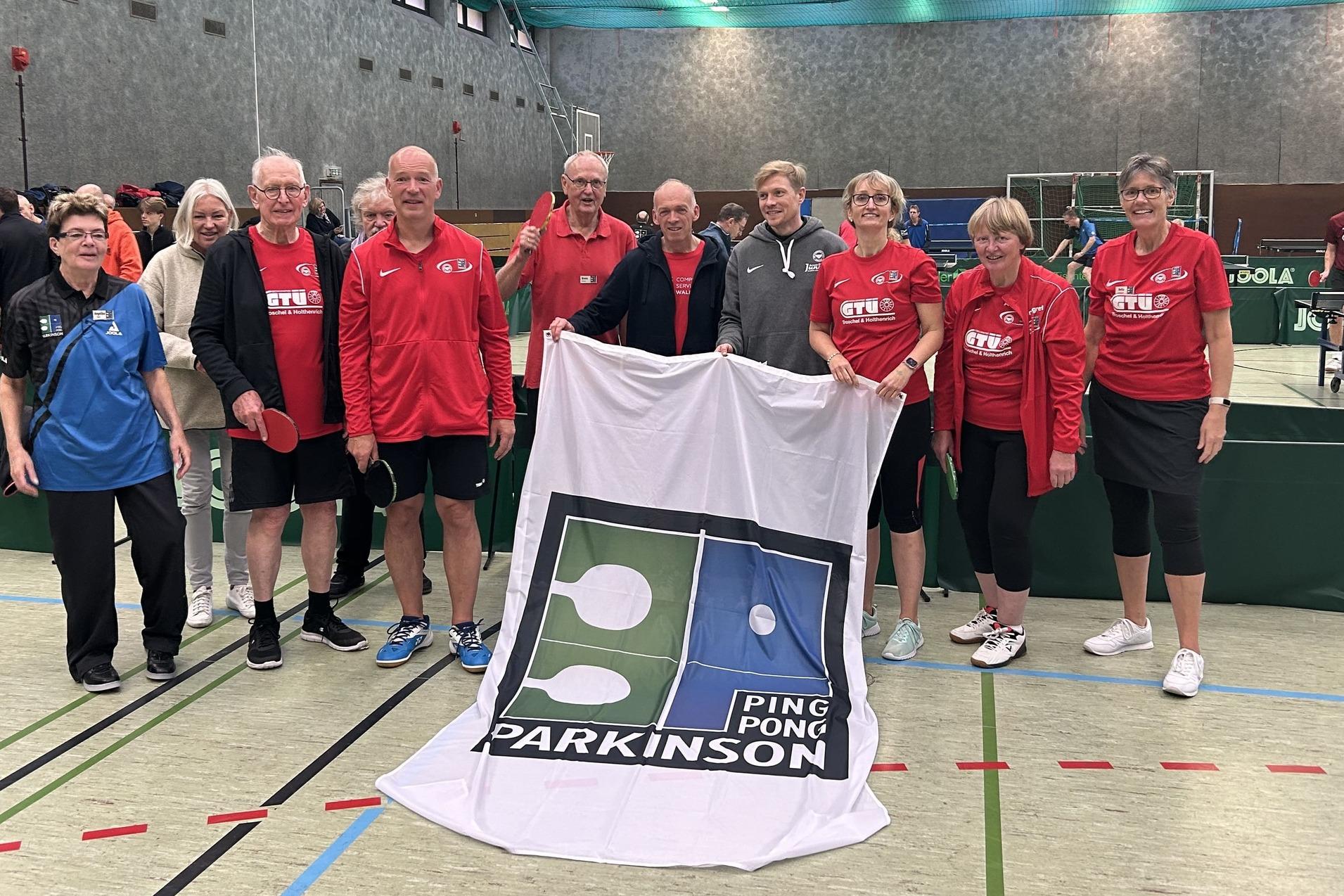 Gruppe von Tischtennisspielern in rot und blau mit Banner "Ping Pong Parkinson" in einer Sporthalle.