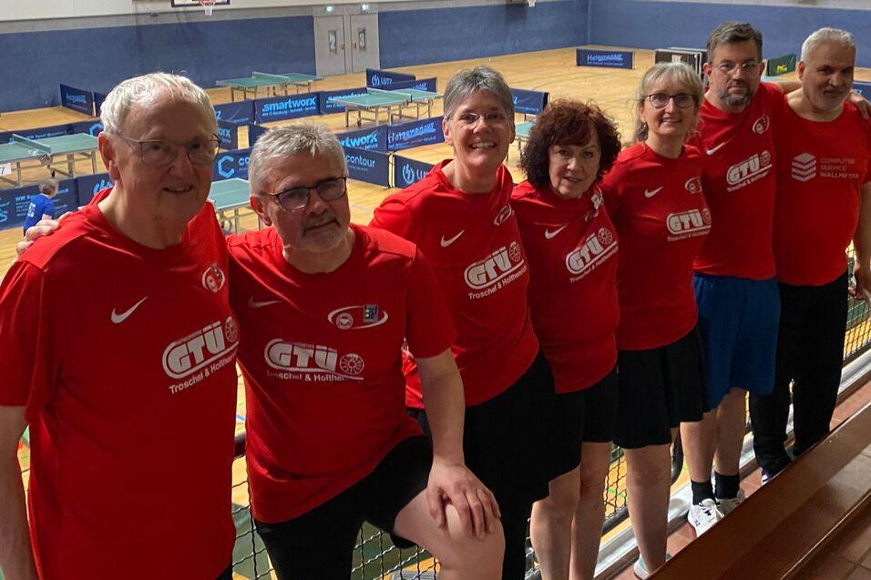 Gruppe von sieben Tischtennisspielern in roten T-Shirts l&auml;chelt auf einer Trib&uuml;ne in einer Sporthalle.