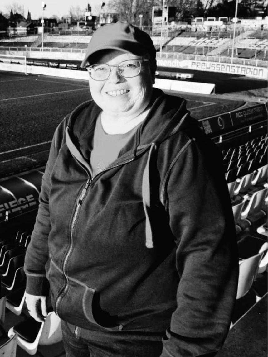 Lächelnde Frau in einem schwarzen Kapuzenpullover steht auf der Tribüne eines Fußballstadions.