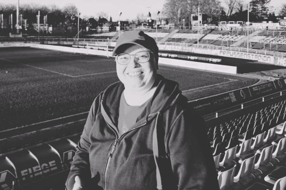 L&auml;chelnder Mensch in Sportkleidung steht auf der Trib&uuml;ne eines leeren Fu&szlig;ballstadions im Schwarzwei&szlig;format.
