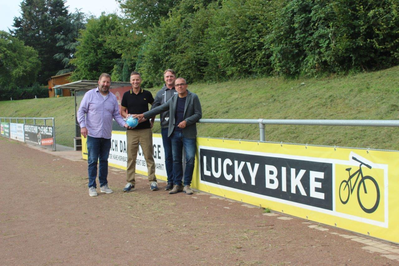 Vier M&auml;nner stehen an einem Sportplatz vor einem Banner mit der Aufschrift &bdquo;LUCKY BIKE&ldquo; und halten einen blauen Ball.