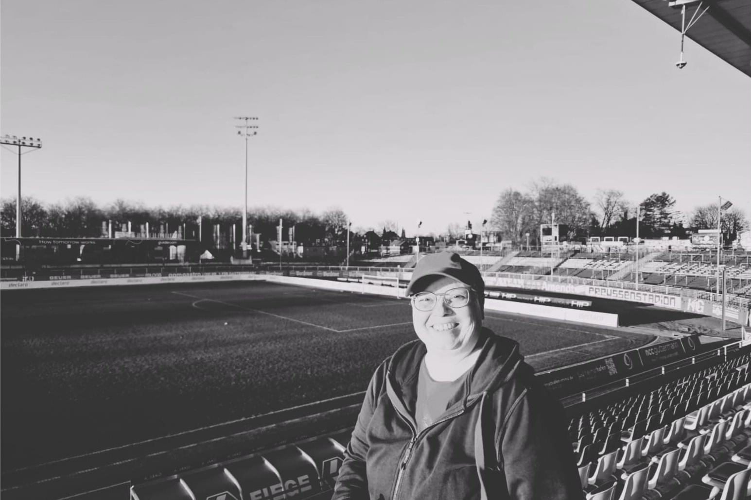 L&auml;chelnder Mensch in Sportkleidung steht auf der Trib&uuml;ne eines leeren Fu&szlig;ballstadions im Schwarzwei&szlig;format.