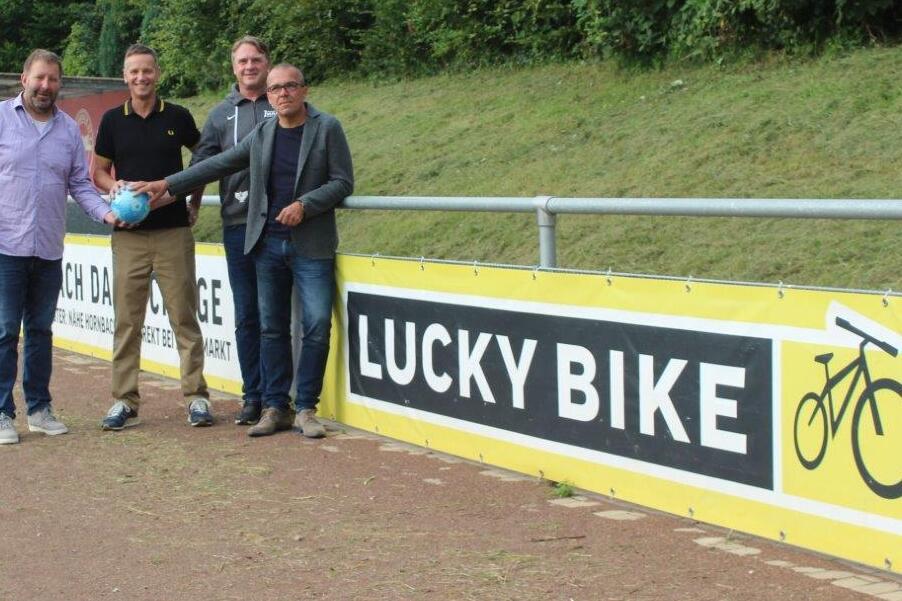 Vier Männer stehen an einem Sportplatz vor einem Banner mit der Aufschrift „LUCKY BIKE“ und halten einen blauen Ball.