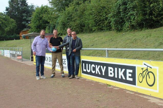 Vier M&auml;nner stehen an einem Sportplatz vor einem Banner mit der Aufschrift &bdquo;LUCKY BIKE&ldquo; und halten einen blauen Ball.