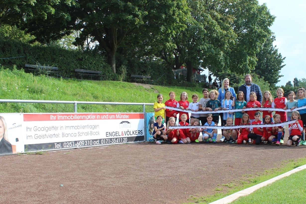 Gruppenfoto von jungen Fu&szlig;ballspielern in Vereinskleidung, posierend vor einem Werbebanner auf dem Sportplatz.