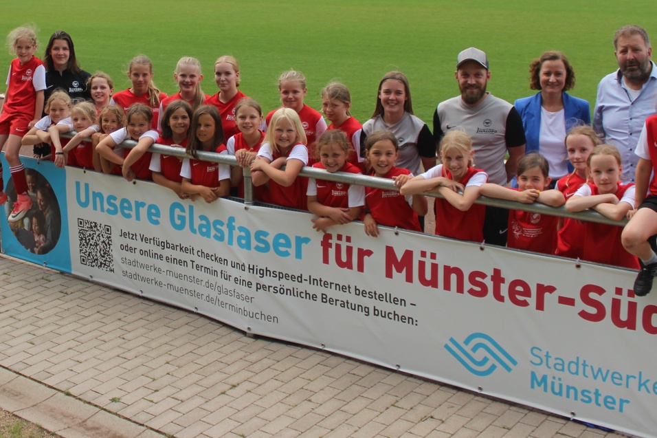 Gruppenshot von M&auml;dchenmannschaft in roten Trikots, posierend am Zaun eines Fu&szlig;ballfeldes mit einem Werbebanner.