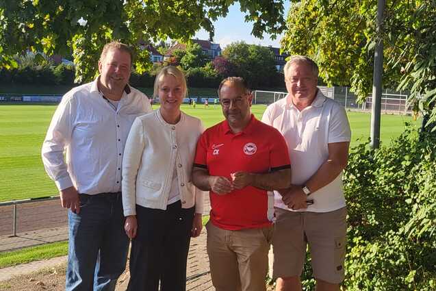 Vier Personen stehen l&auml;chelnd vor einem Fu&szlig;ballfeld, umgeben von B&auml;umen, in einem sonnigen Stadionumfeld.