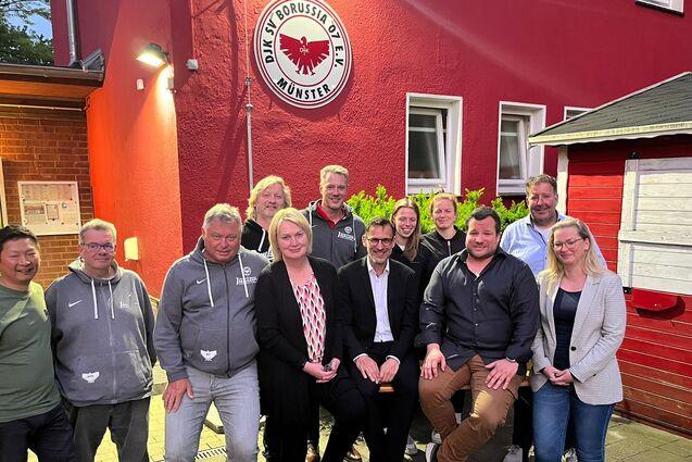 Gruppenzusammenkunft vor der Vereinsfassade von Borussia M&uuml;nster, Mitglieder posieren l&auml;chelnd in zwei Reihen.