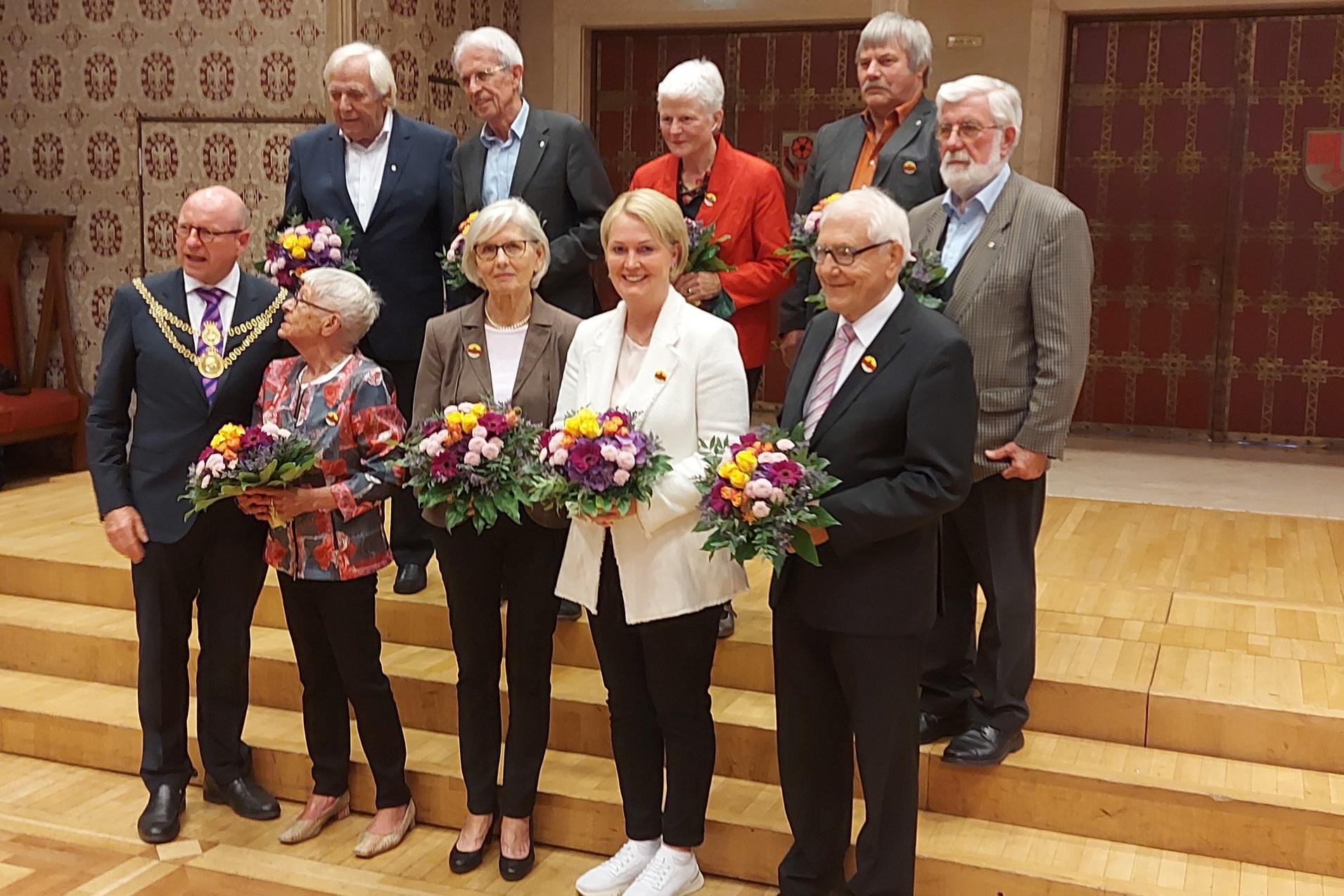 Gruppe von Personen mit Blumensträußen auf einer Bühne in einem festlichen Raum.