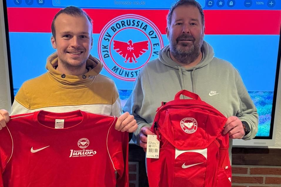 Zwei M&auml;nner halten ein rotes T-Shirt und einen roten Rucksack vor einem Bildschirm mit dem Logo von Borussia M&uuml;nster.