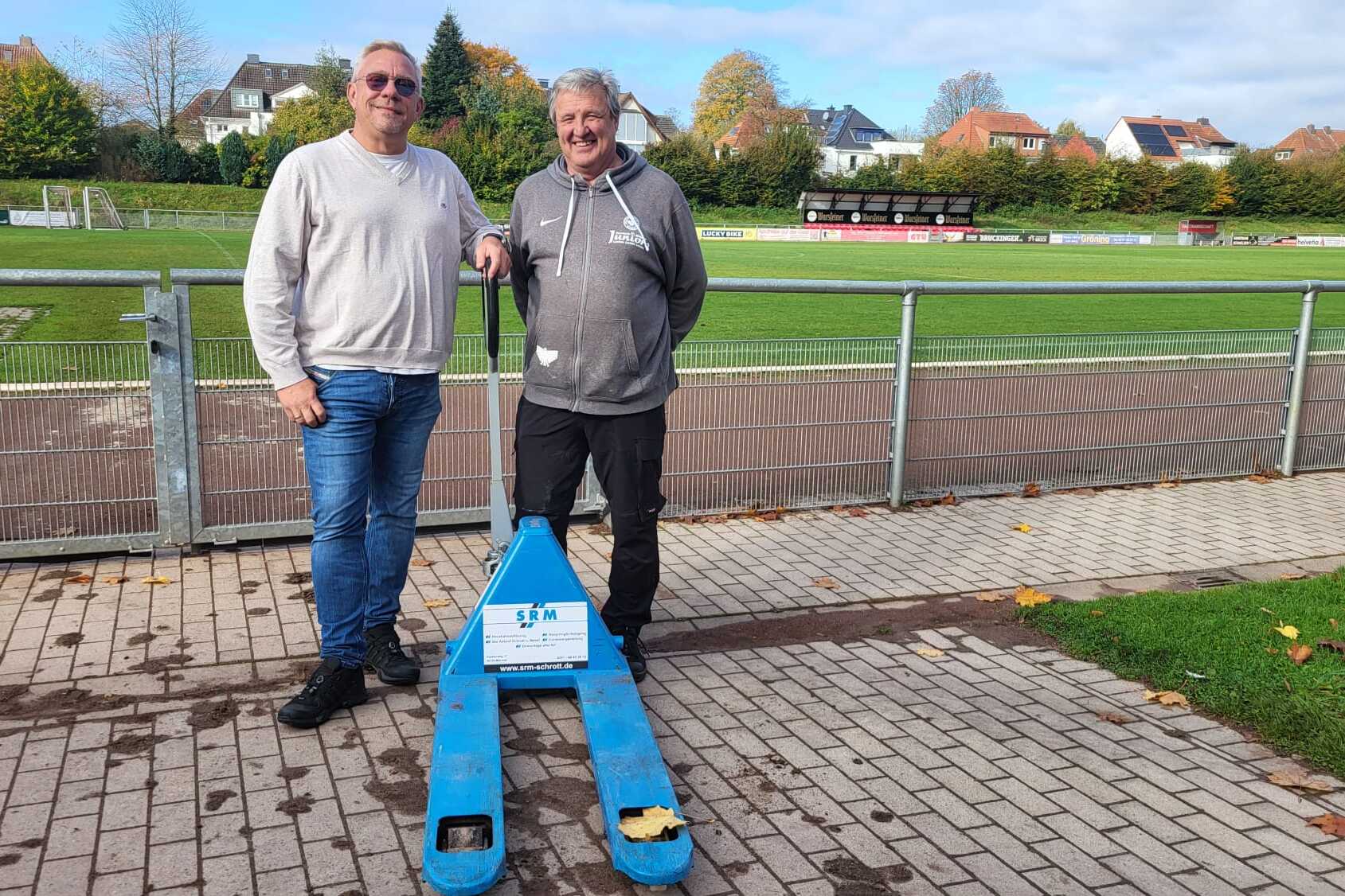 Zwei M&auml;nner stehen im Freien neben einem blauen Sportger&auml;t auf einem Fu&szlig;ballplatz mit Trib&uuml;ne im Hintergrund.