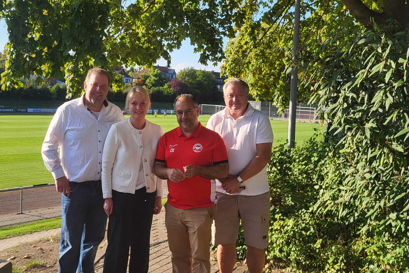 Vier Personen stehen l&auml;chelnd vor einem Fu&szlig;ballfeld, umgeben von B&auml;umen, in einem sonnigen Stadionumfeld.