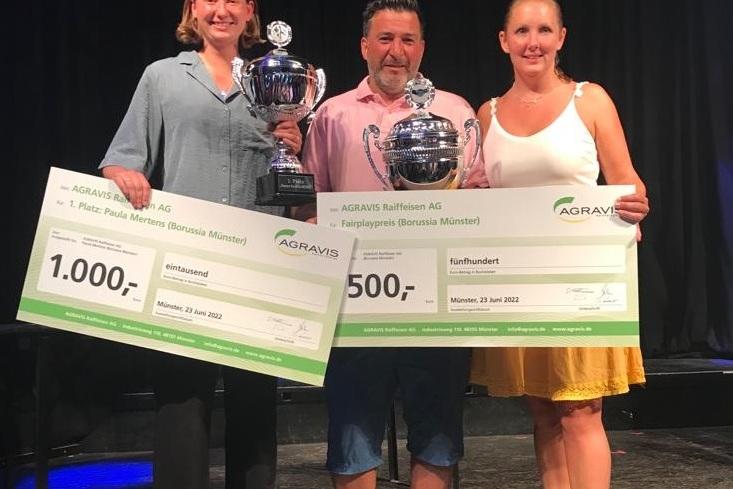 Drei Personen halten Pokale und Schecks f&uuml;r die Platzierungen bei einer Sportveranstaltung auf einer B&uuml;hne.